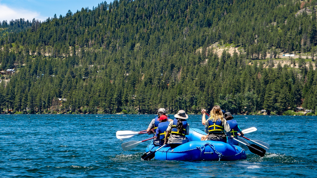 A raft with people rowing it moves across Donner Lake with a forested mountain in the background.